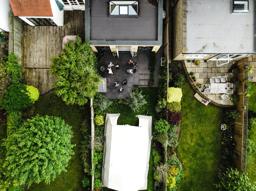 An aerial view of a well-maintained garden showing a variety of lush green plants, trees, and a neatly trimmed lawn. In the center, there is a paved patio area with several people gathered, engaging i